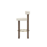 Modern luxury bar chair with cream upholstery, double-roll backrest, walnut legs, and brass-finished footrest, designed by Nolita Harbour. Side view