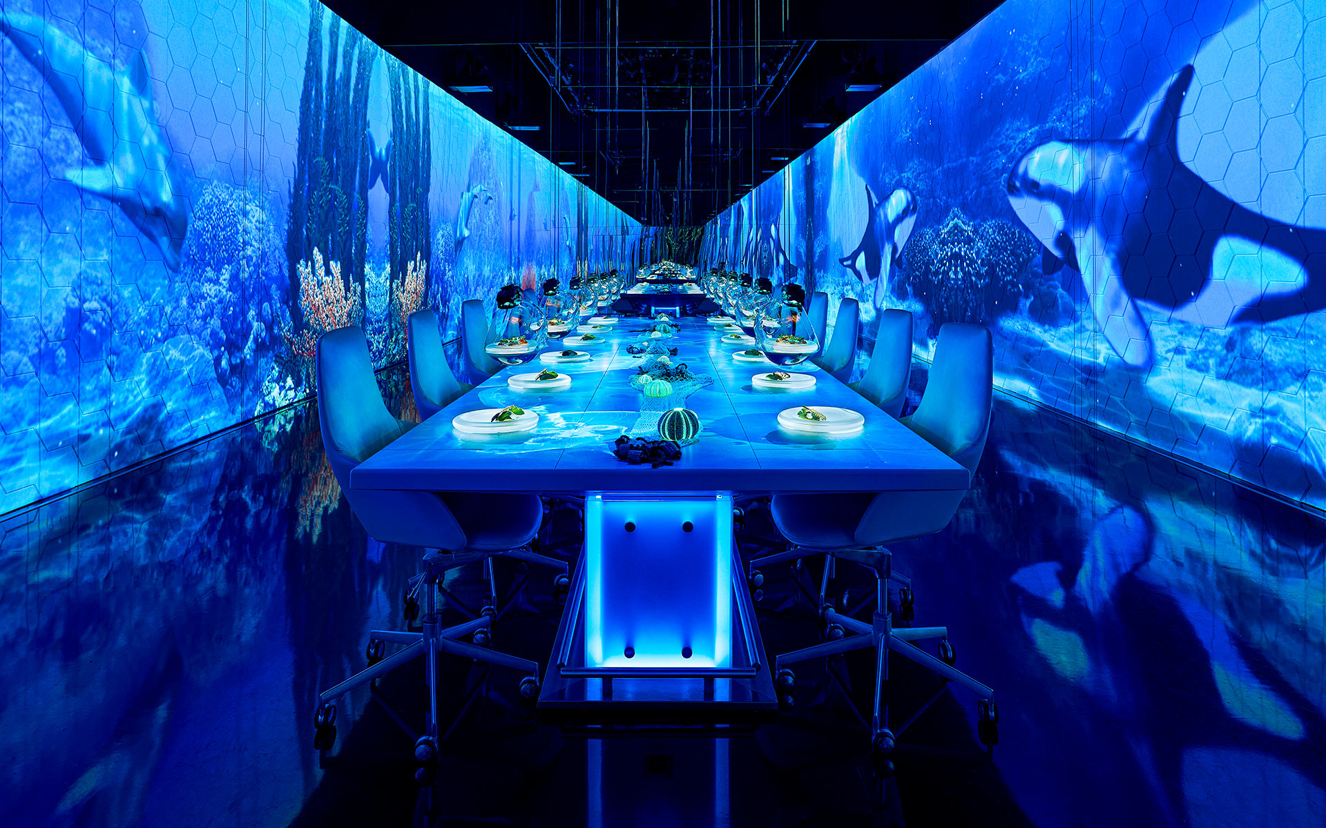 This image showcases a futuristic, immersive dining experience with a high-tech, underwater theme. The walls and ceiling are covered in large-scale digital projections of marine life, including whales and coral reefs, creating the illusion of being submerged in the ocean. The long, sleek dining table is illuminated with blue lighting, enhancing the deep-sea ambiance. The setting combines fine dining with digital artistry, offering a multi-sensory experience that merges gastronomy with technology. - Nolita Harbour 