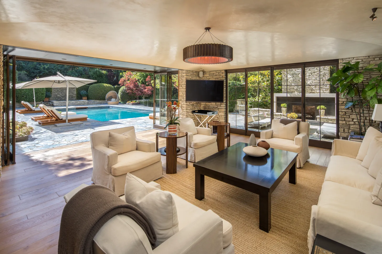  A cozy and elegant living room with white sofas, a dark wooden coffee table, and a rustic stone fireplace. The living room seamlessly connects to an outdoor pool area with lush greenery and sun loungers, thanks to large glass doors. - Nolita Harbour