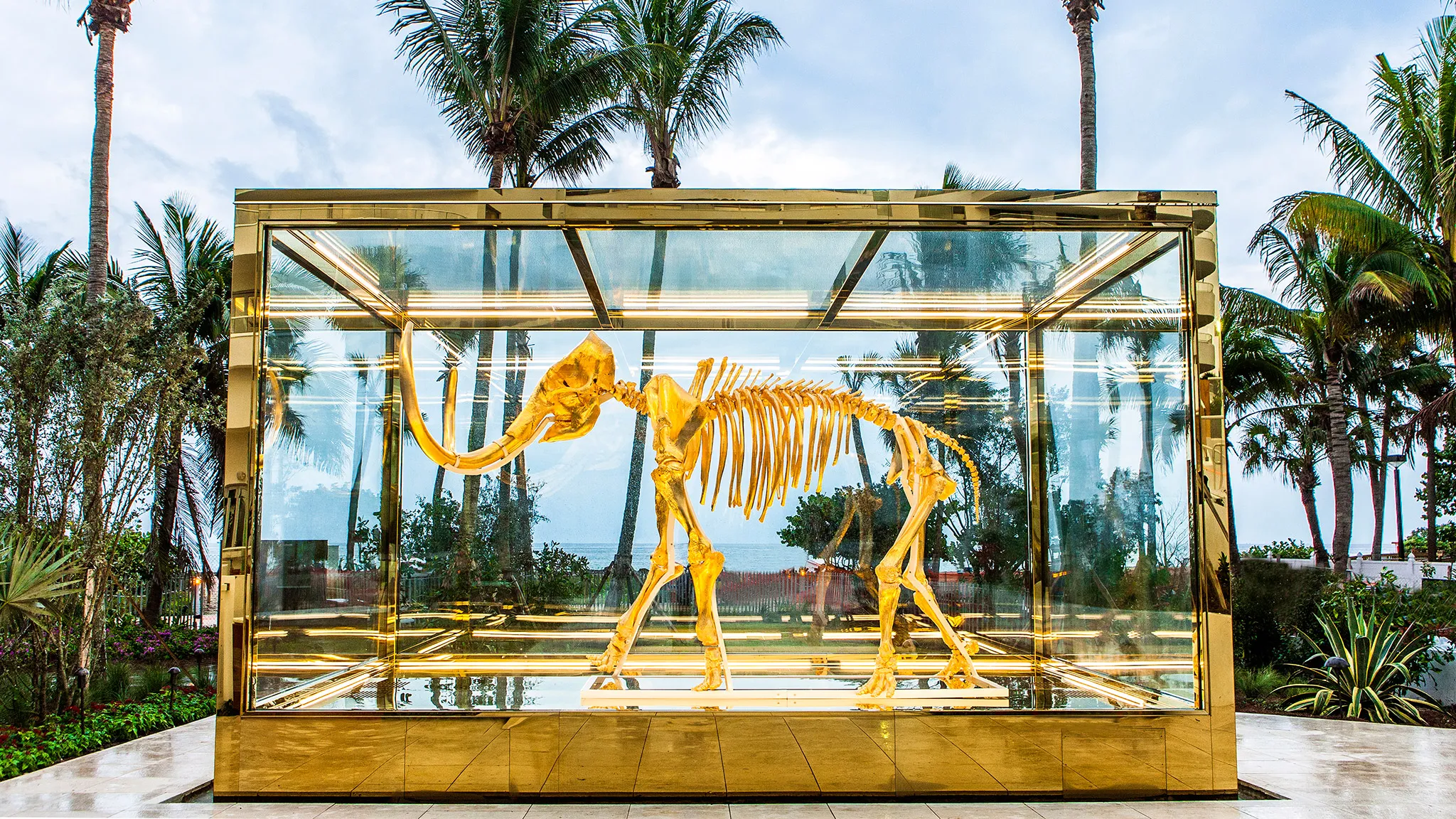 A striking art installation featuring a gold-plated mammoth skeleton encased in a glass and gold frame. Set against a tropical backdrop with palm trees and an ocean view, the display combines luxury and nature, creating a visually stunning contrast. Nolita Harbour 