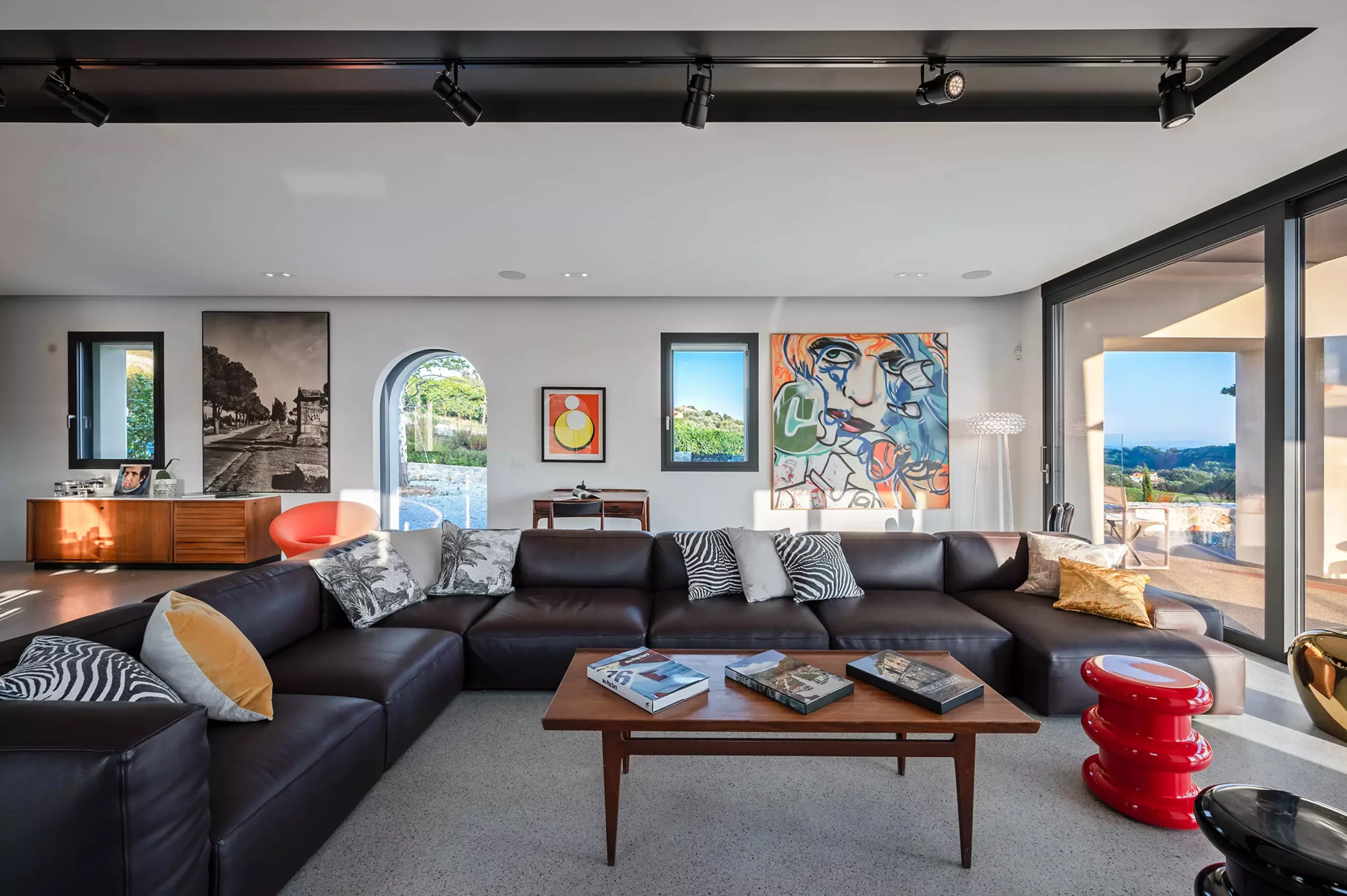 A modern living room featuring a large dark brown leather sectional sofa, a wooden coffee table, a bold red sculptural side table, and a mix of black-and-white and colorful abstract artwork. Large glass doors provide natural light and scenic outdoor views. - Nolita Harbour