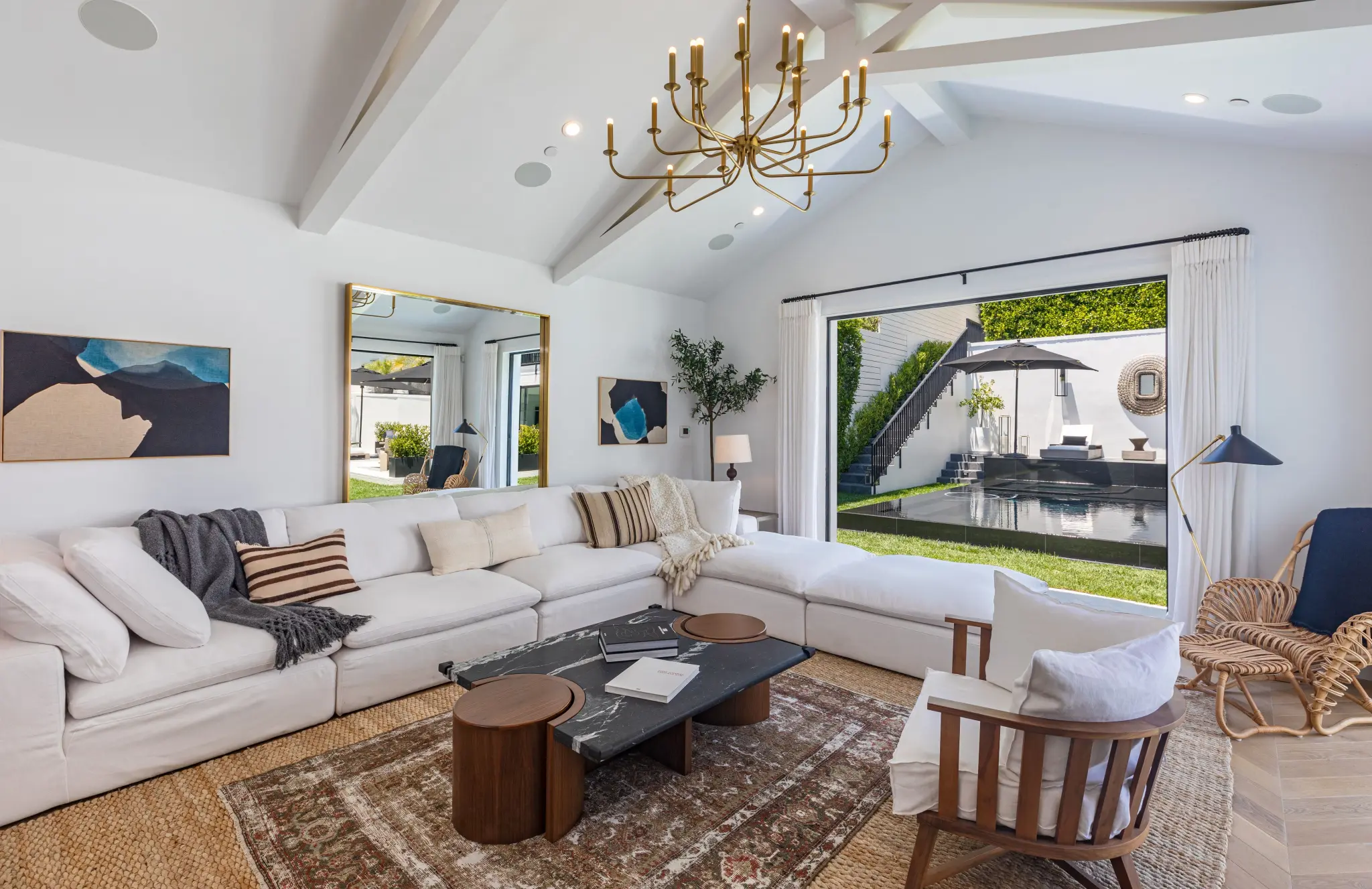 A bright and airy living room with a vaulted ceiling, a white sectional sofa, a large gold chandelier, and a black marble coffee table. The space opens to a backyard featuring a pool and modern outdoor seating. - Nolita Harbour