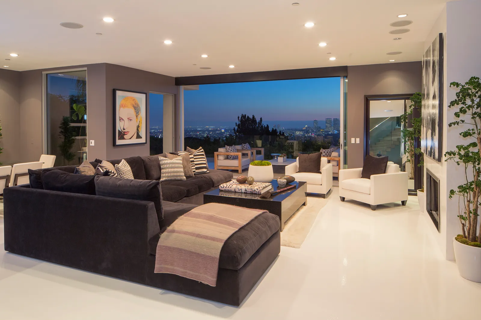 A modern, high-end living room with dark-toned decor, a black sectional sofa, white accent chairs, and a large wall-mounted artwork. The space has floor-to-ceiling windows offering a breathtaking city skyline view at dusk. - Nolita Harbour