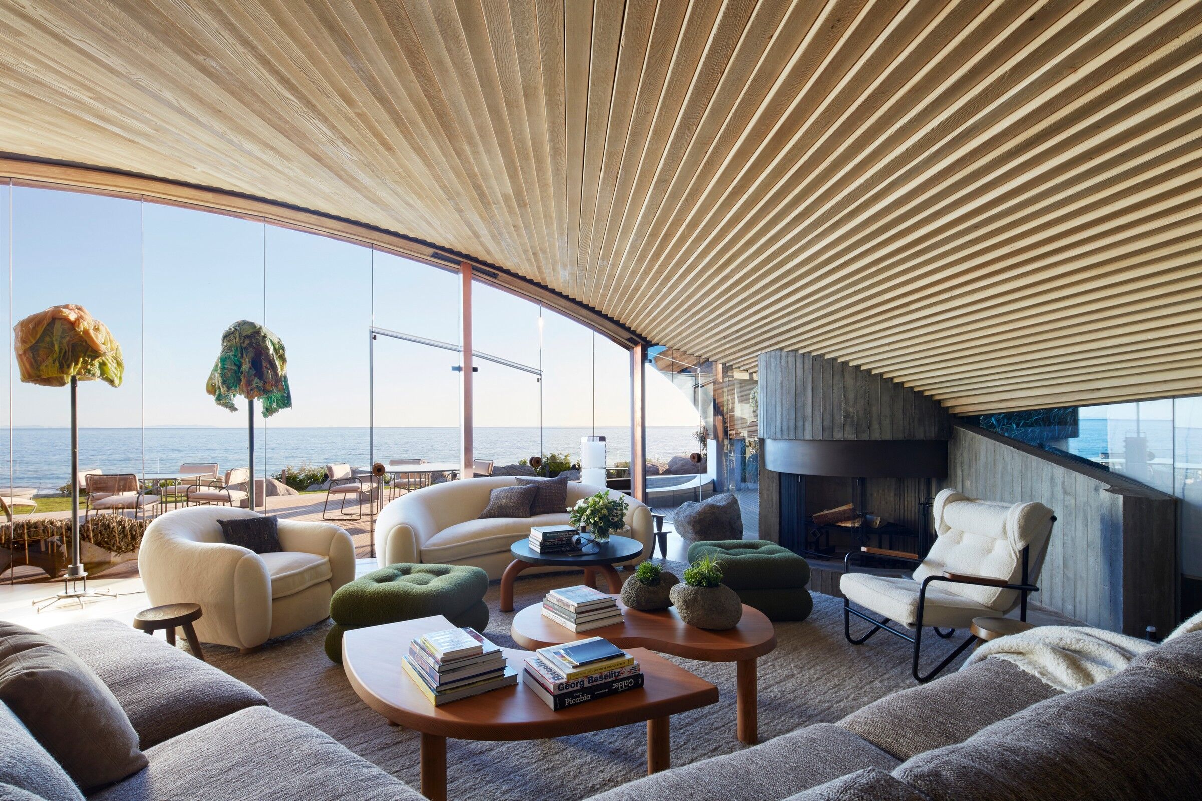 A modern coastal living space with a curved wooden ceiling, floor-to-ceiling glass walls, and panoramic ocean views. The room features plush, neutral-toned seating, organic-shaped wooden coffee tables, and green ottomans. A black fireplace adds warmth, while outdoor seating seamlessly extends the space to the waterfront. Natural textures and earthy tones create a harmonious, relaxing ambiance. - Nolita Harbour 