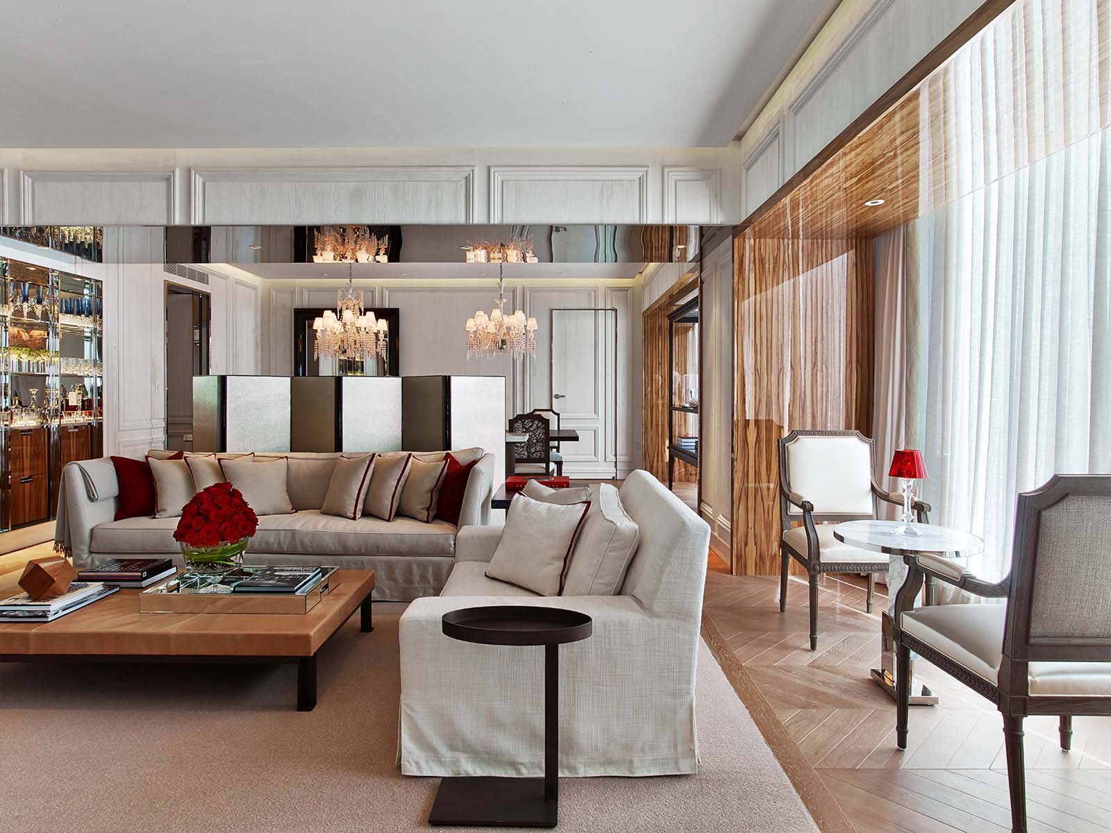 A luxury hotel living room with elegant décor, featuring plush seating, a mirrored wall, chandeliers, a bar area, and floor-to-ceiling sheer curtains that enhance the space with natural light. The neutral color palette is accented with red details for a sophisticated touch. - Nolita Harbour 