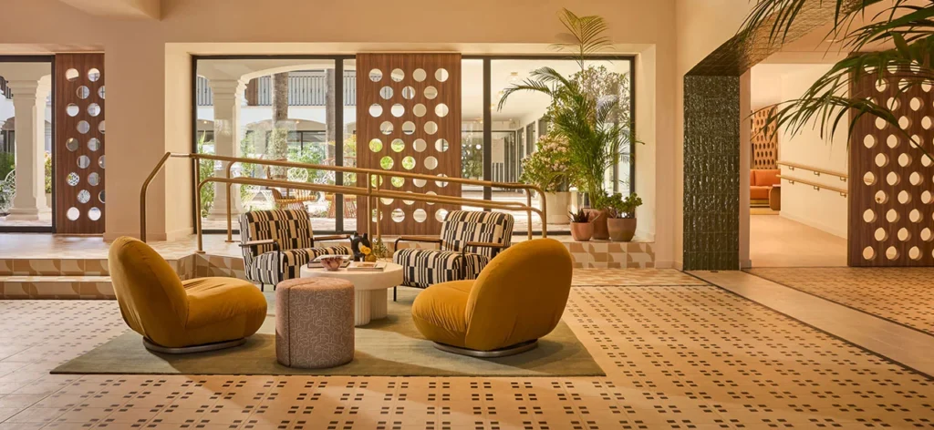 Stylish Kimpton Los Monteros hotel lounge with modern armchairs, patterned seating, indoor plants, and large windows overlooking a Mediterranean courtyard.