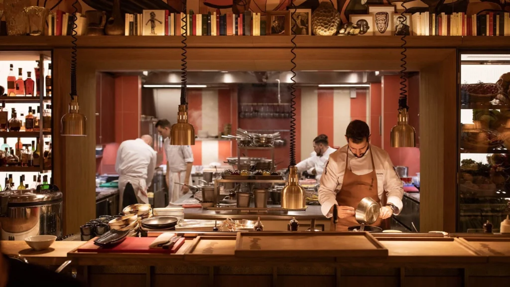 Open kitchen in a luxury fine dining restaurant, showcasing chefs at work behind a bespoke wood counter, warm brass lighting, and curated shelving that blends culinary ritual with refined restaurant interior design.