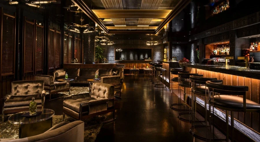 Moody luxury cocktail bar interior with plush seating, dark wood paneling, brass accents, and layered ambient lighting, illustrating modern fine dining design trends focused on intimacy and opulence.