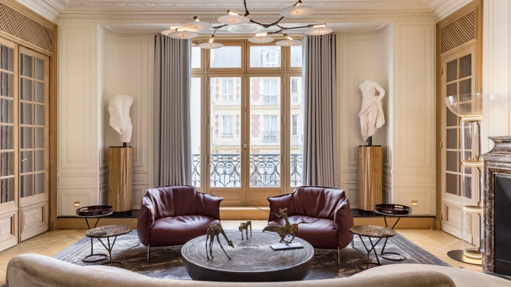 Refined Parisian apartment living room featuring restored Haussmann moldings, tall French windows, sculptural contemporary seating, and a balanced dialogue between classical architecture and modern urban interior design.