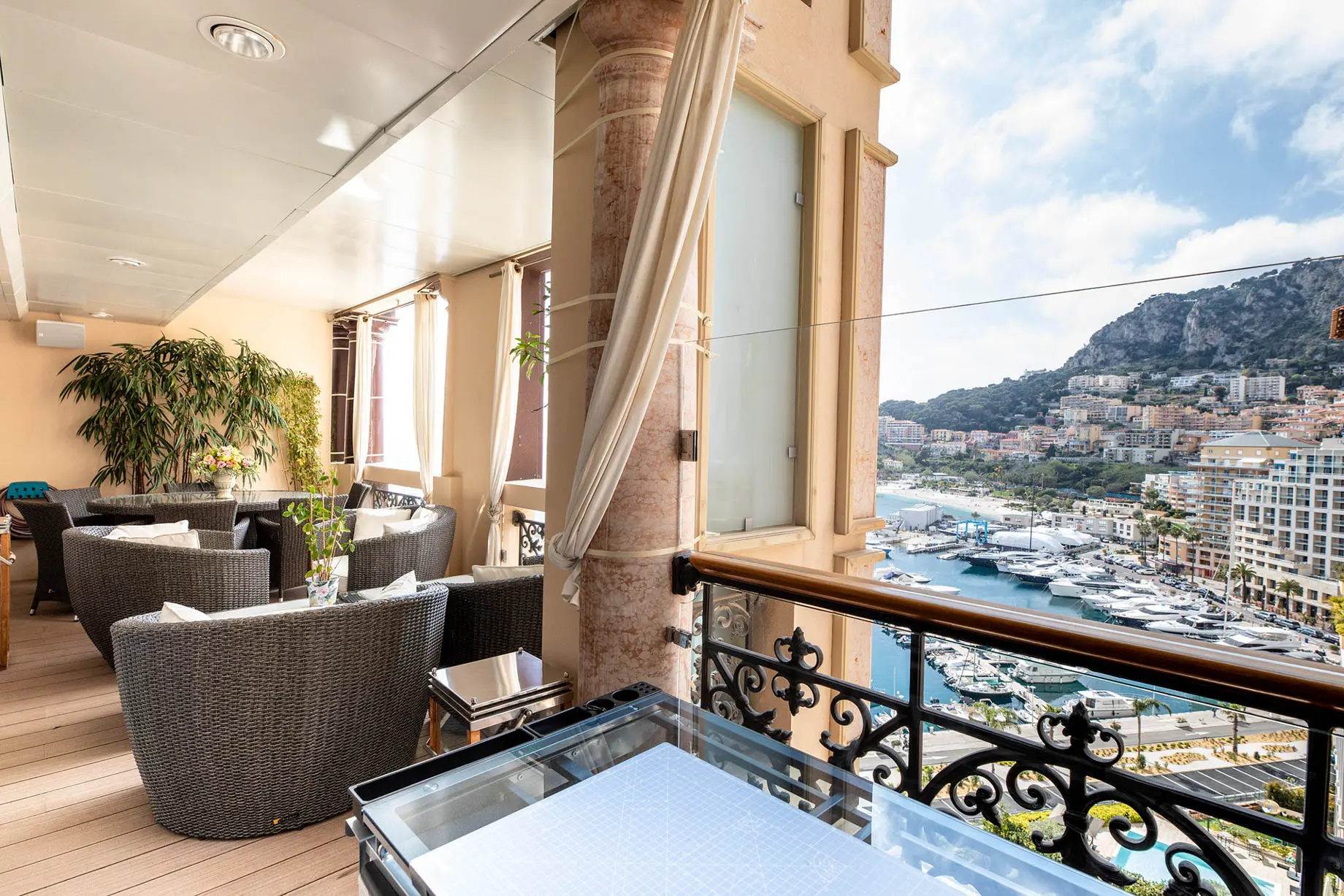 Sophisticated terrace with wicker furniture and plants overlooking a vibrant marina and hillside cityscape under a clear blue sky, typical of Monaco penthouses.