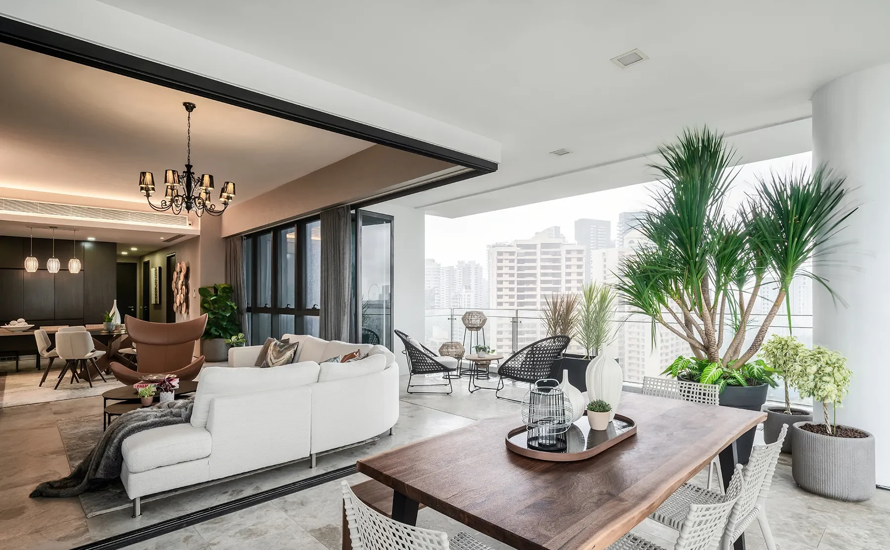 A modern open-concept living space with a patio. Features include a white sofa, dining area, chandelier, large plants, and city skyline views, creating a spacious, airy feel.