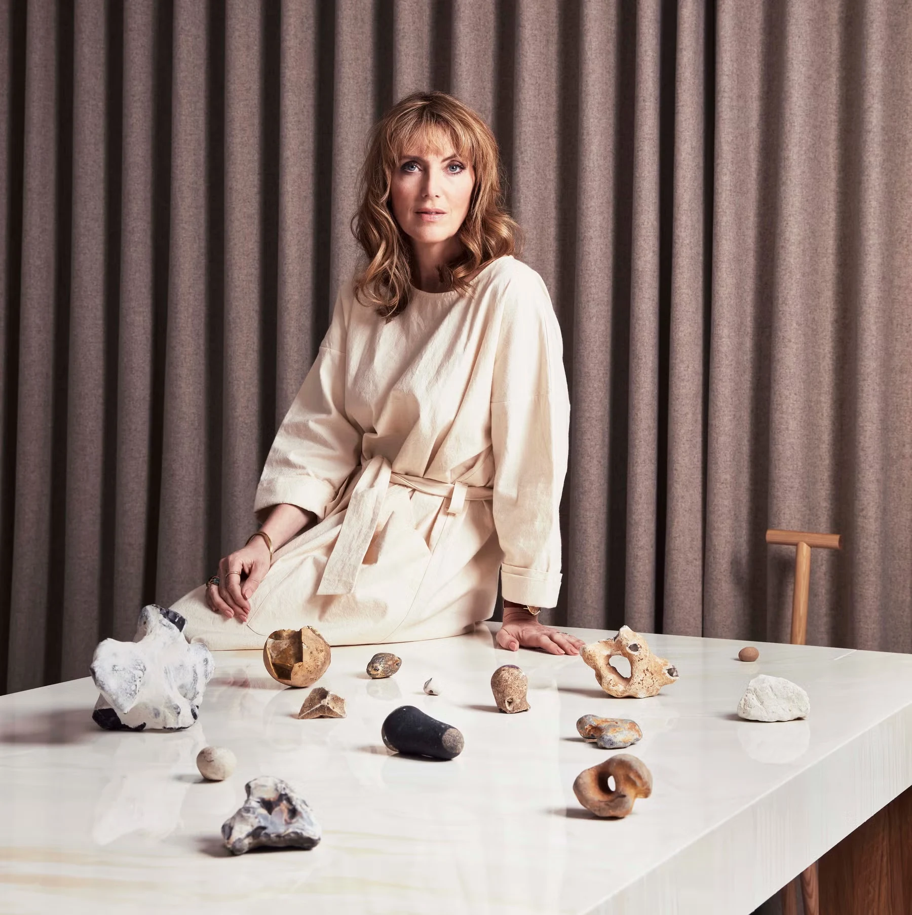 Faye Toogood in a cream dress sits on a table with various rocks and fossils. The background curtain is taupe, creating a calm and contemplative atmosphere.