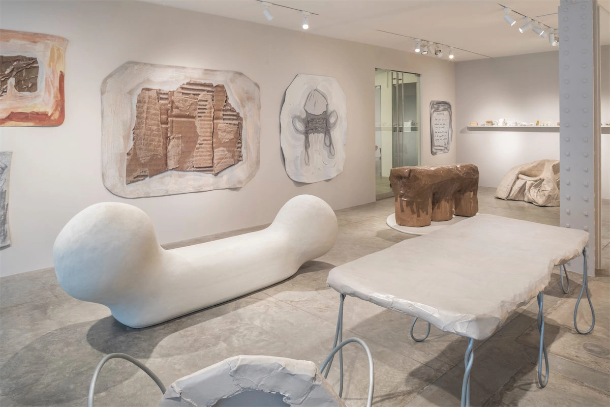 Art gallery with abstract sculptures and textured wall art. Features a bone-shaped bench, organic forms, and muted tones, creating a modern, contemplative atmosphere.