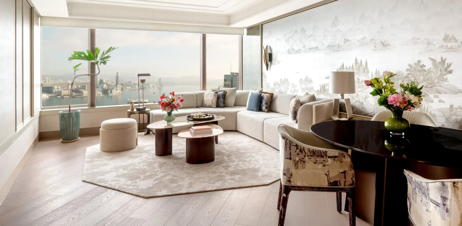 Luxurious, sunlit living room with large windows showing a city view. Features a plush sectional sofa, elegant decor, floral arrangements, and a round dining table.