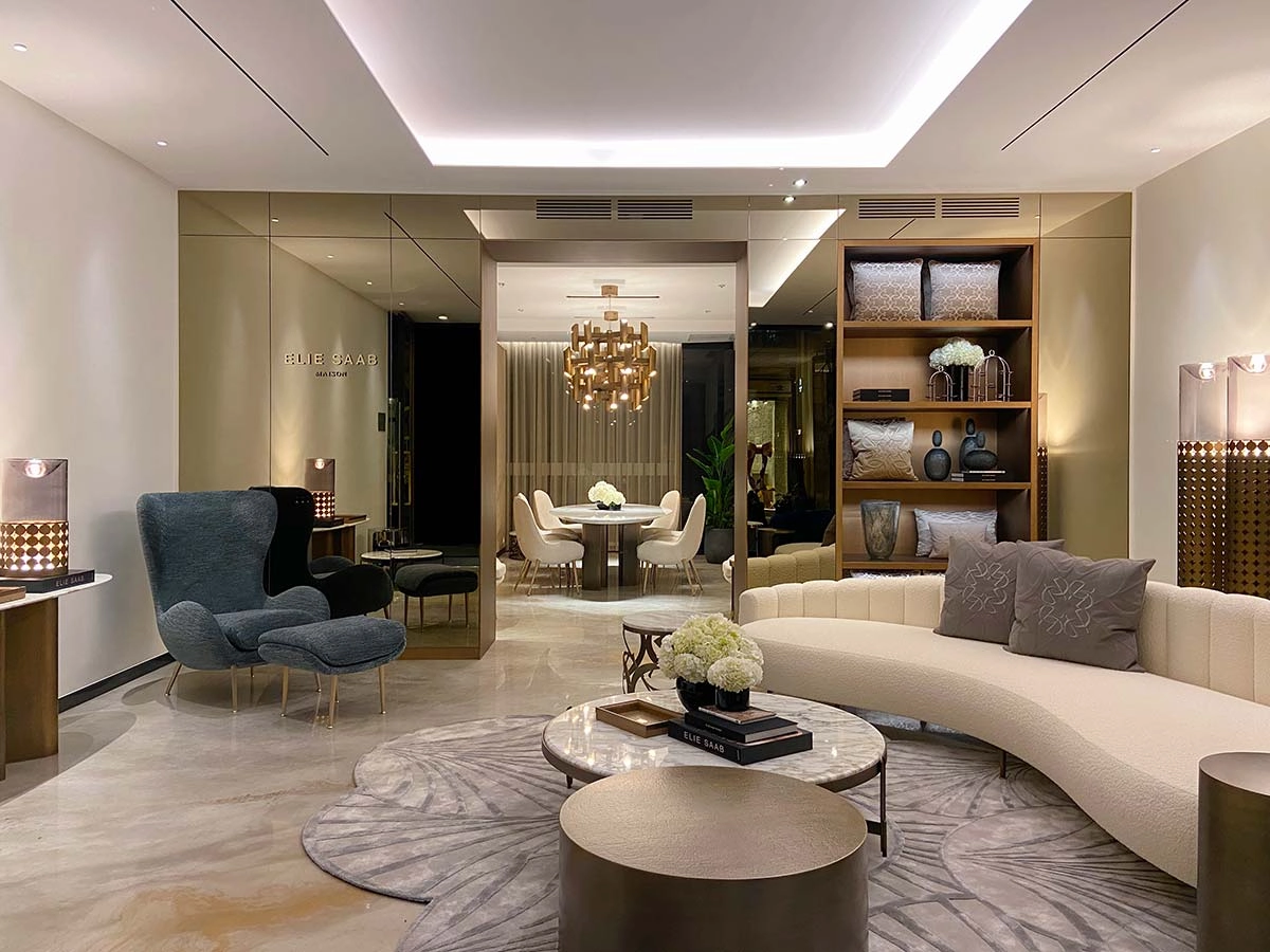 Luxurious modern Elie Saab Maison living room featuring a curved cream sofa, marble tables, a tall armchair, and elegant lighting. Shelves with decor and a glimpse into a dining area with a chandelier create a sophisticated ambiance.