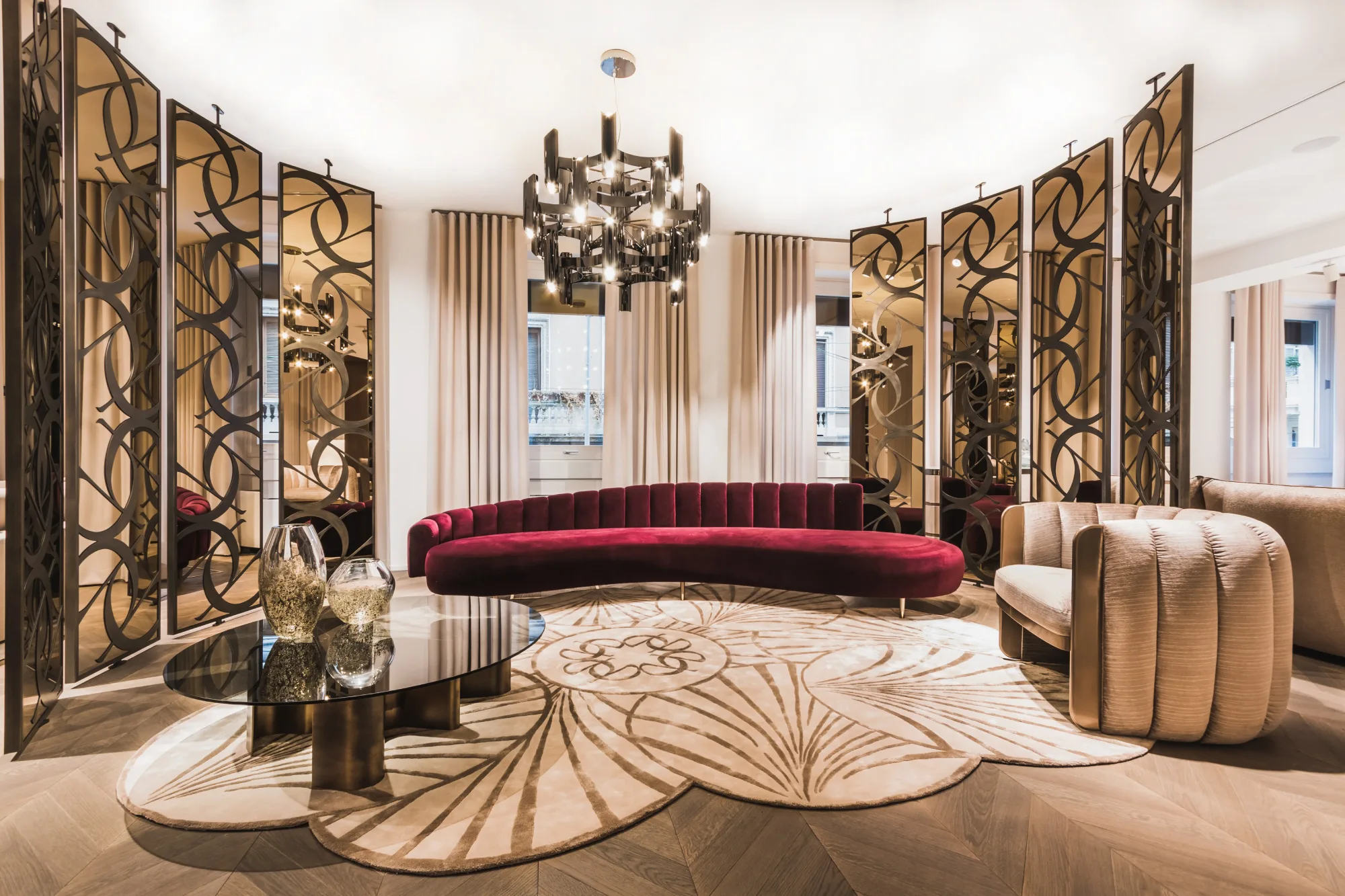 Upscale Elie Saab Maison living room with an elegant chandelier, curved red sofa, patterned rug, and modern screen dividers, exuding a luxurious and warm ambiance.