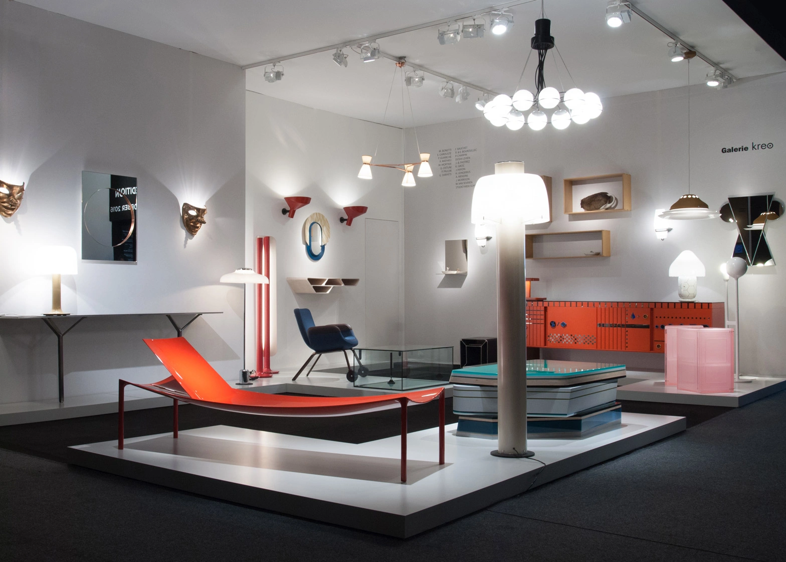 Exhibition room with modern furniture and lighting, featuring a red chaise lounge, eclectic lamps, wall-mounted shelves, and vibrant decor accents.