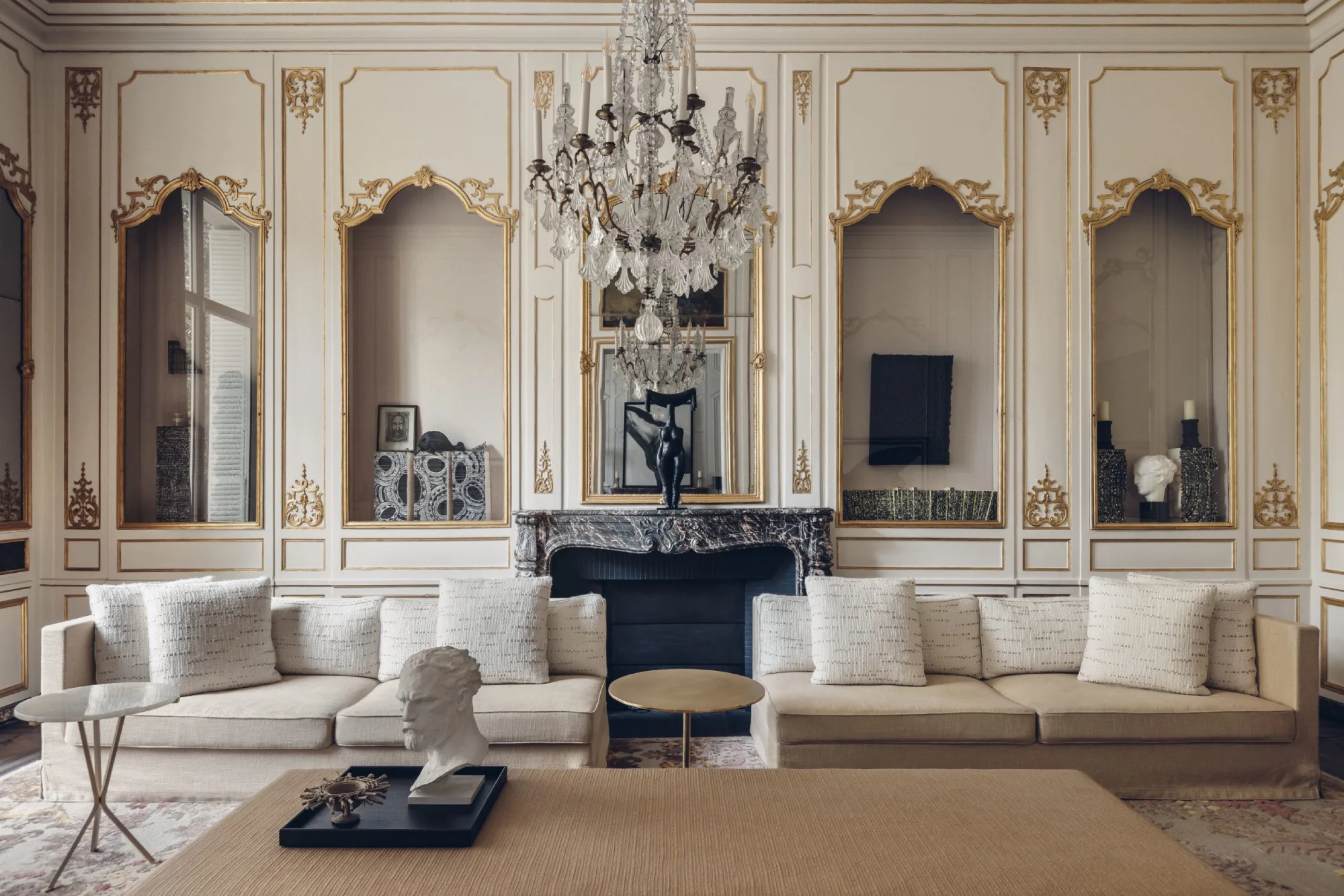 Elegant living room with a cream sectional sofa, ornate gold-trimmed white walls, a grand crystal chandelier, and a marble fireplace, exuding sophistication.
