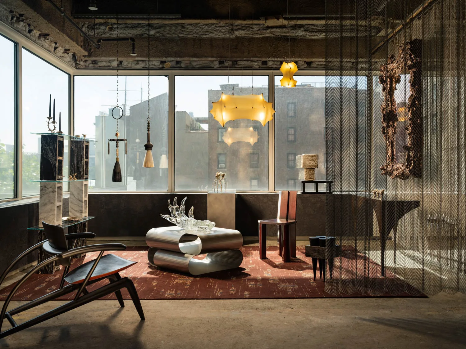 A modern room with industrial decor features large windows, unique hanging lights, a metallic coffee table with glass sculptures, and chic furniture on a patterned rug.