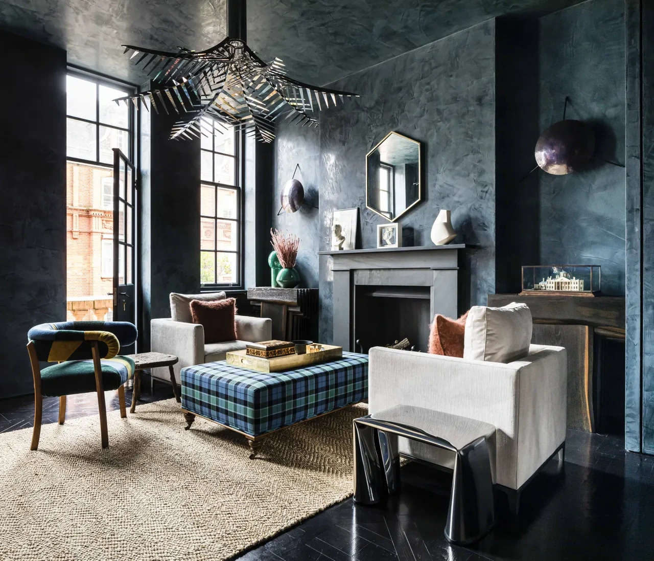 Sophisticated living room with dark walls, modern chandelier, plaid ottoman, white chairs, and a fireplace. A cozy, contemporary ambiance.