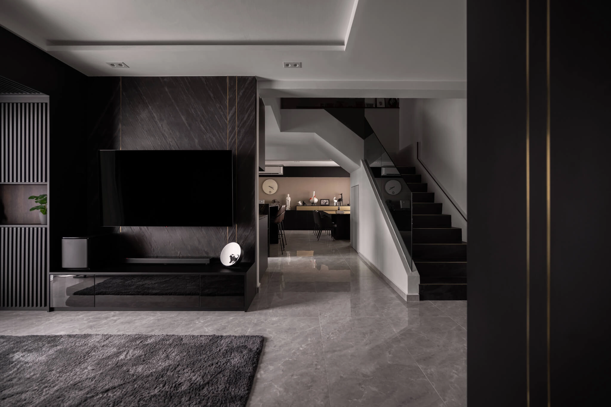 Sleek modern living room with dark tones, large TV on black accent wall, glossy marble floors, a staircase, and a kitchen in the background.