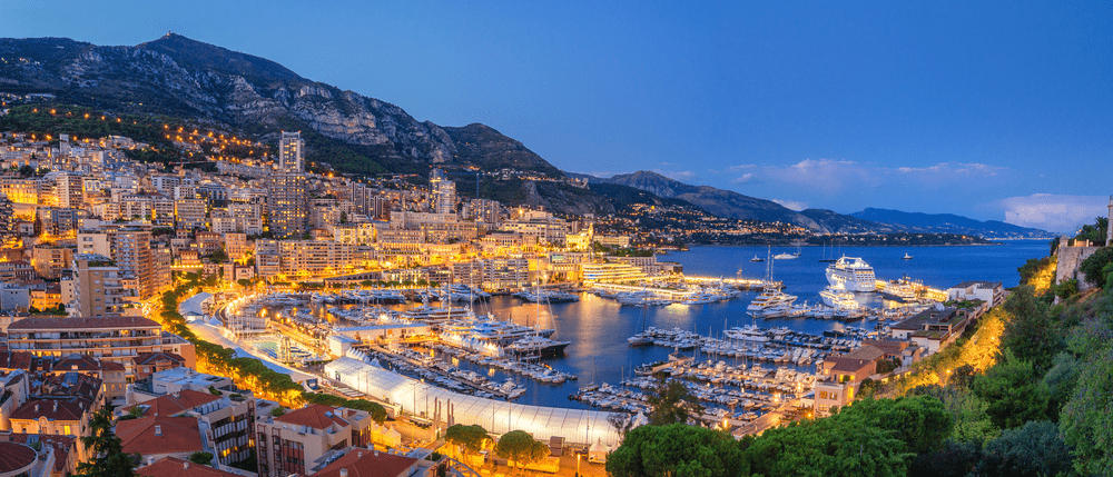 Aerial view of a vibrant harbor at dusk, with luxury yachts docked. City lights illuminate the scene, reflecting on the water, surrounded by mountains.