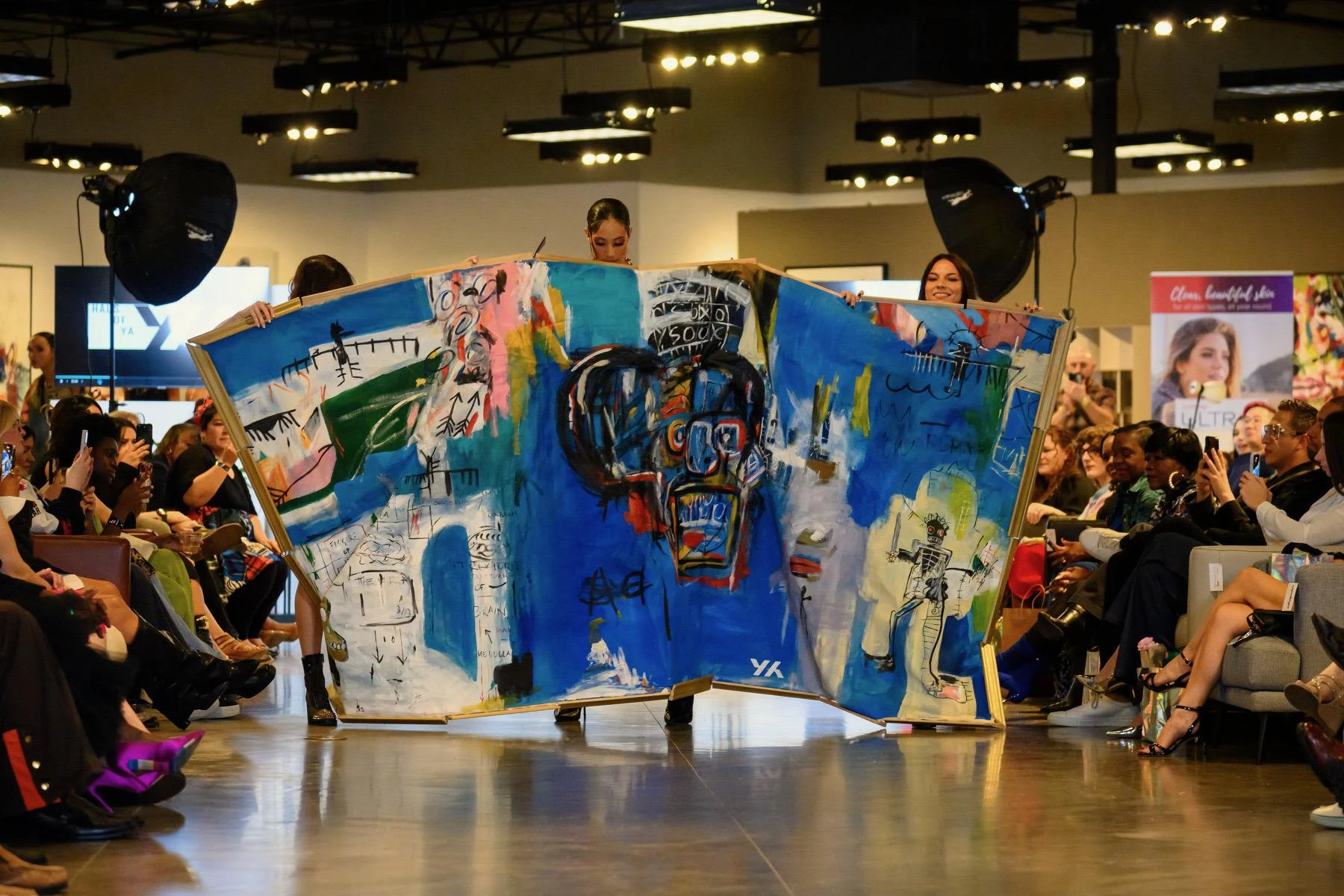 A vibrant artwork held by models on a runway features bold colors and abstract shapes on canvas. The audience watches attentively, capturing the energetic mood.