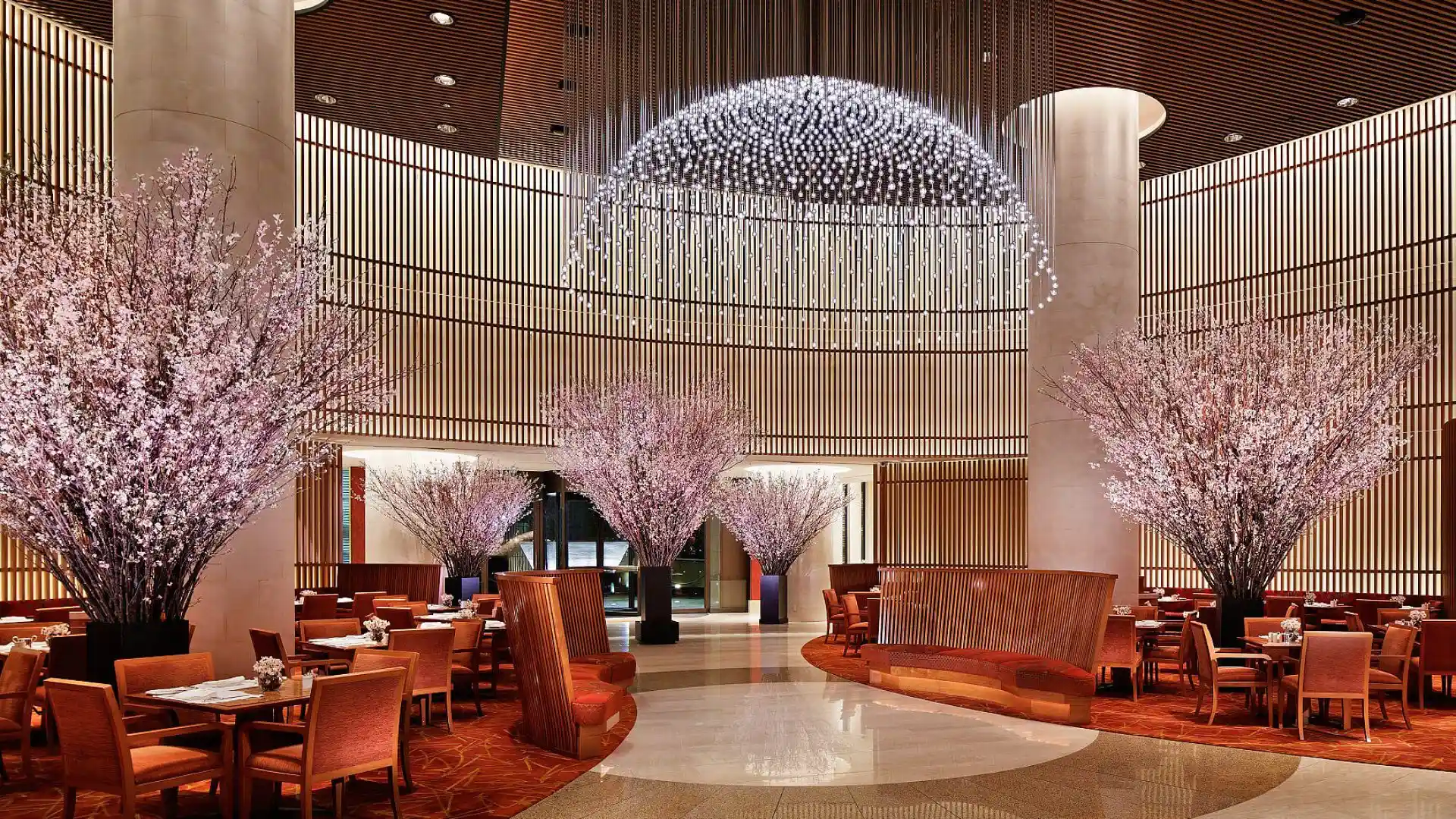 Luxurious restaurant interior with elegant cherry blossom trees, warm lighting, round tables, and a stunning dome chandelier, creating a serene ambiance.