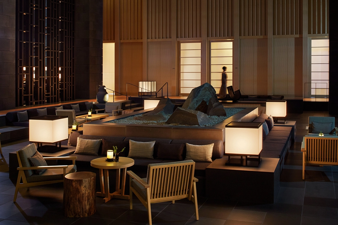 A serene luxury hotel lounge with dim lighting, featuring a central rock garden surrounded by cozy seating. Warm wooden accents and soft lamps create a tranquil ambiance. It demonstrates the comfort and serenity of luxury hotels.