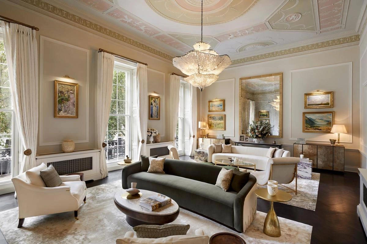 Elegant living room with a high ornate ceiling, large windows, and plush seating. A central chandelier adds luxury. Art and flowers enhance warmth.