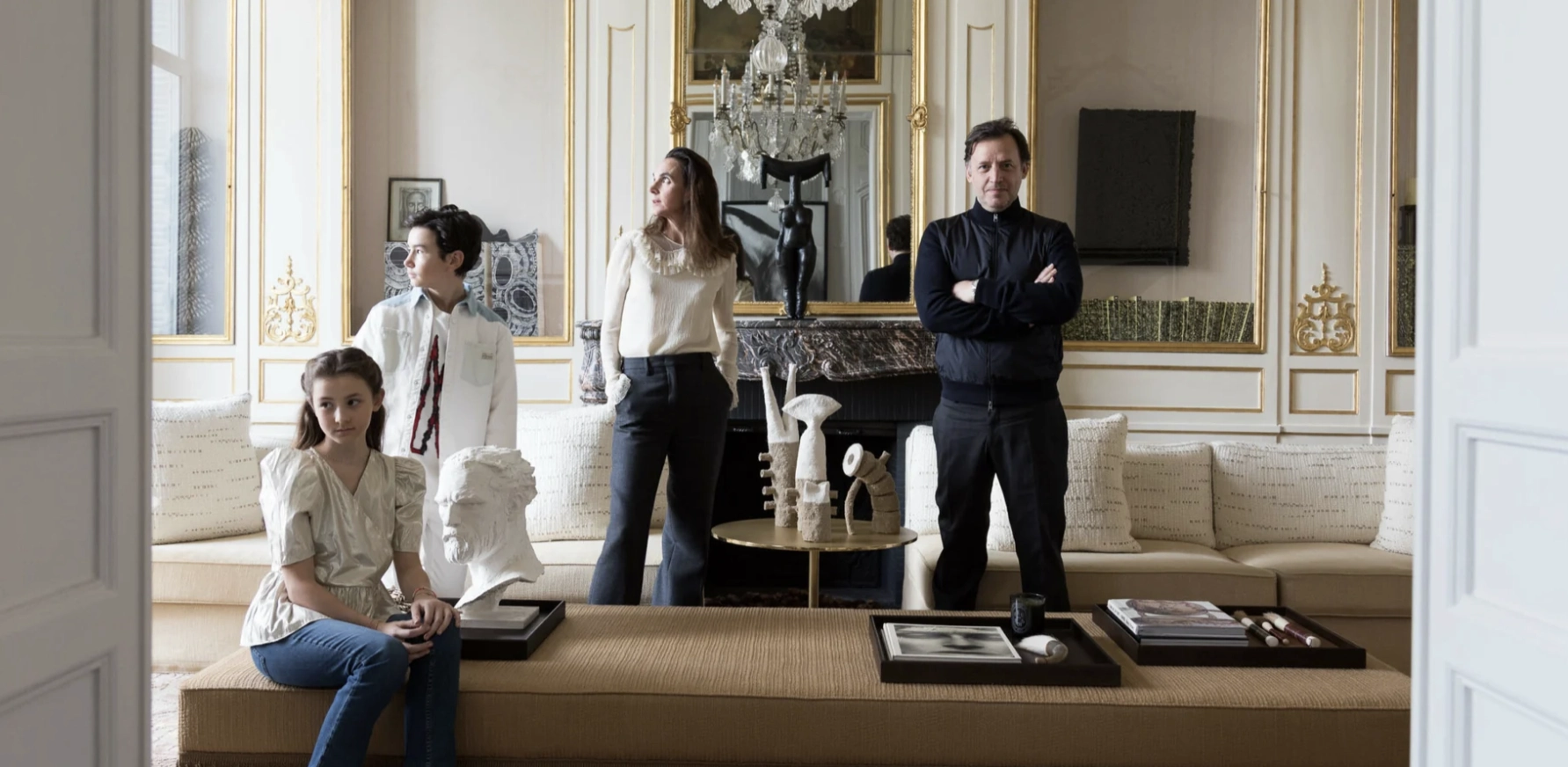 A family of four, Gilles & Boissier and children, stands in an elegant living room with ornate details. They appear relaxed and thoughtful. A marble bust and framed art add sophistication.