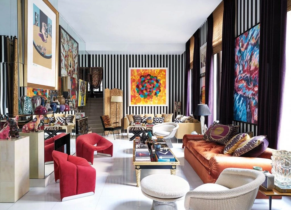 Eclectic living room with vibrant artwork, striped walls, a mix of colorful furniture, including red and orange couches, and plush patterned pillows.