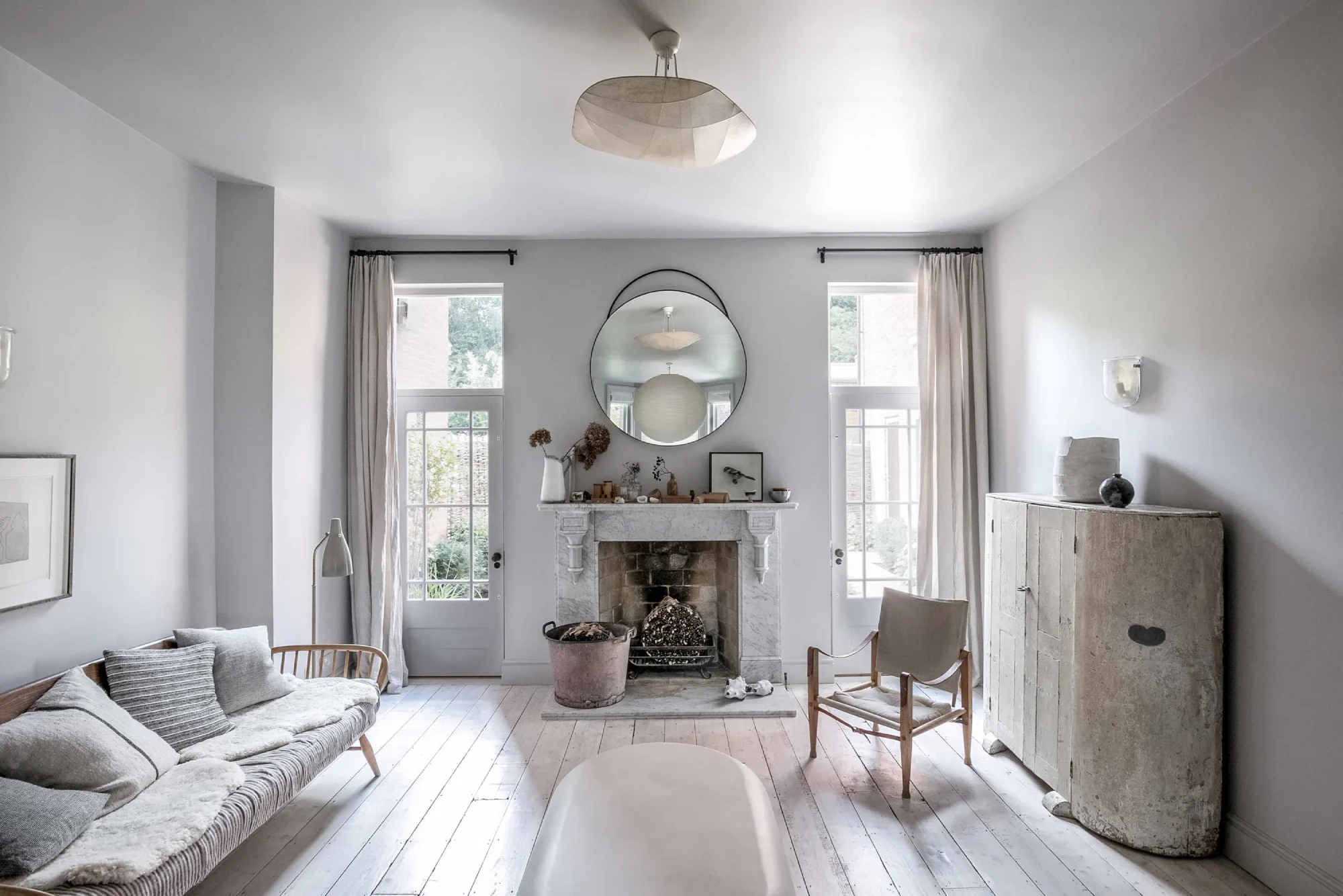 A cozy, minimalist living room with neutral tones. It features a light gray sofa, a rustic fireplace with a round mirror above, and natural light from tall windows.