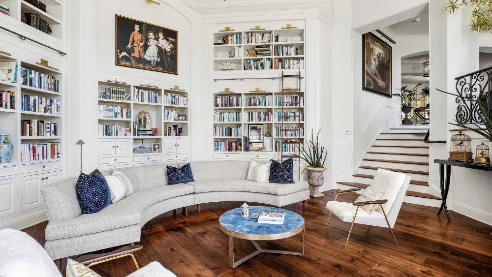 Sophisticated living room with a curved gray sofa, blue cushions, and a round marble table on wooden floors. Shelves with books, art, and stairs in view.