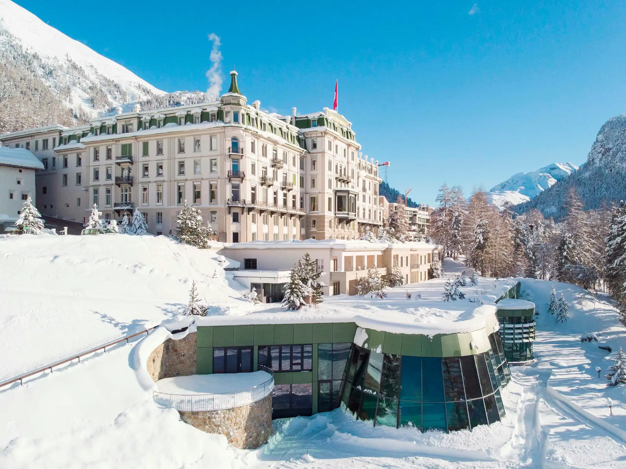 Grand hotel kronenhof, an example of swiss alsp resorts, set against snowy mountains under clear blue skies. The luxurious building is surrounded by snow-covered trees, creating an elegant, serene winter scene.