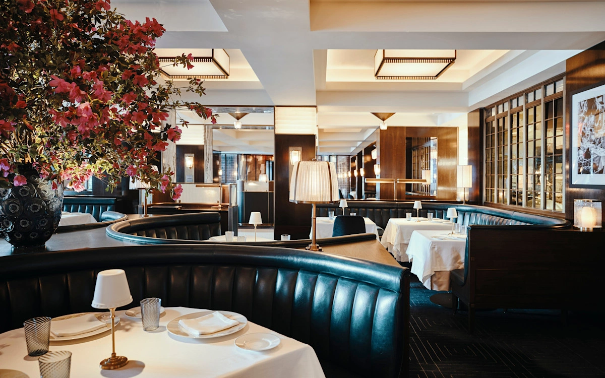 Elegant restaurant interior with leather booths, white tablecloths, soft lighting, and a large vase of pink flowers. The atmosphere is warm and inviting.