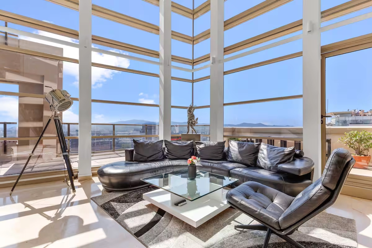 Luxurious modern living room with large corner windows, black leather sectional sofa, glass coffee table, sleek chair, and decorative tripod lamp. Bright and airy ambiance.
