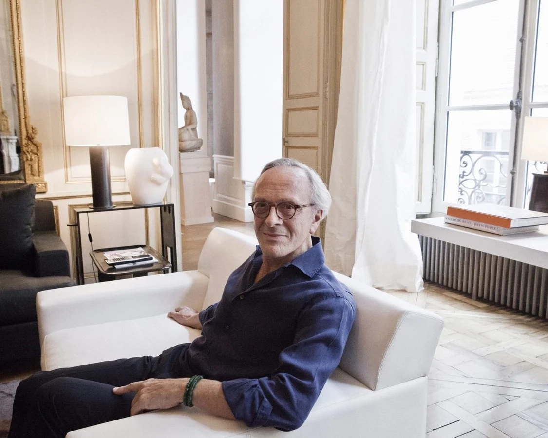 Christian Liaigre sits on a white sofa in an elegant room with large windows, wooden floors, and contemporary sculptures, conveying a calm and sophisticated atmosphere.