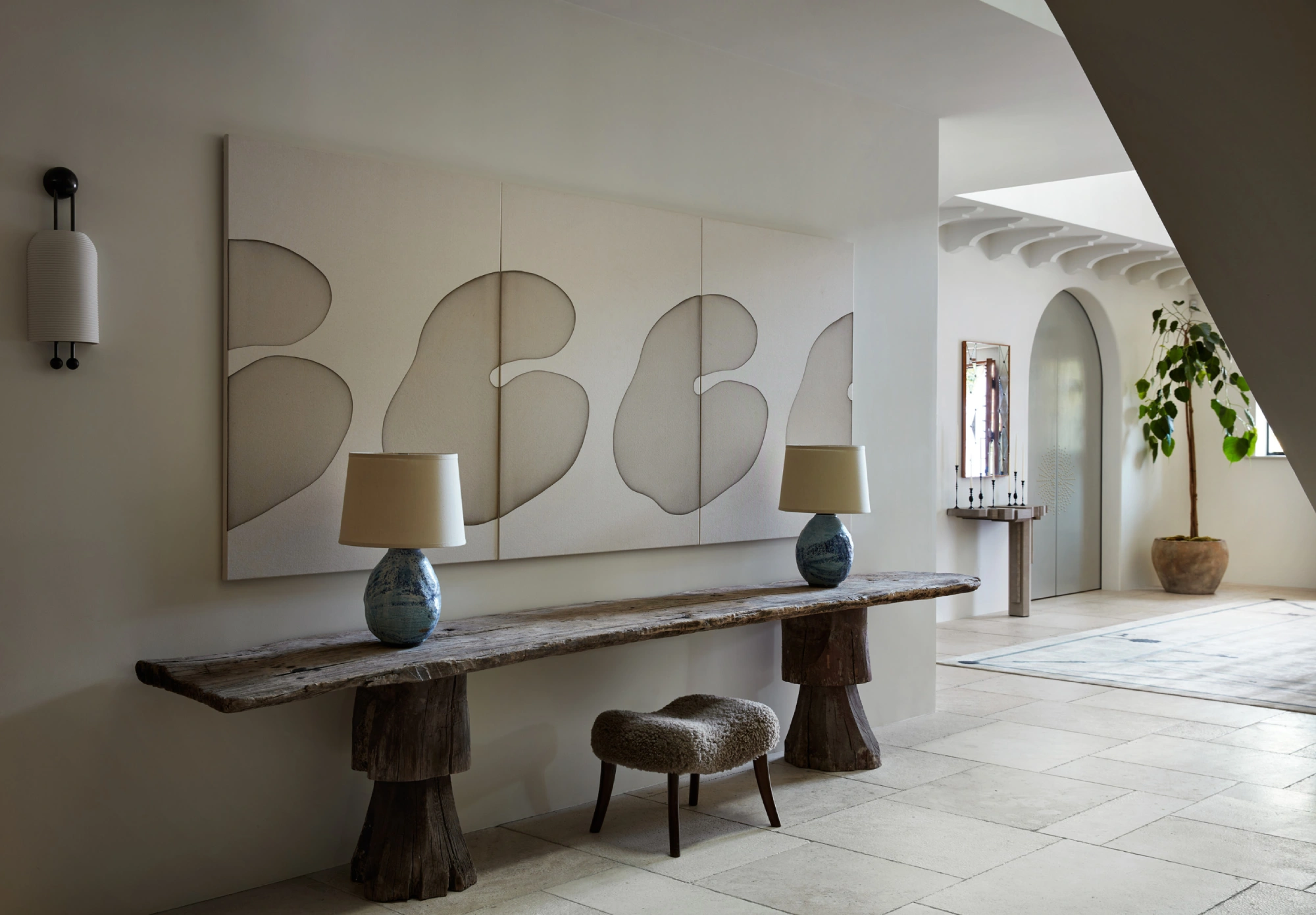 Minimalist hallway designed by Studio Shamshiri with abstract wall art above a rustic wooden console. Two lamps sit on the console. A plush stool on light tiles adds warmth.