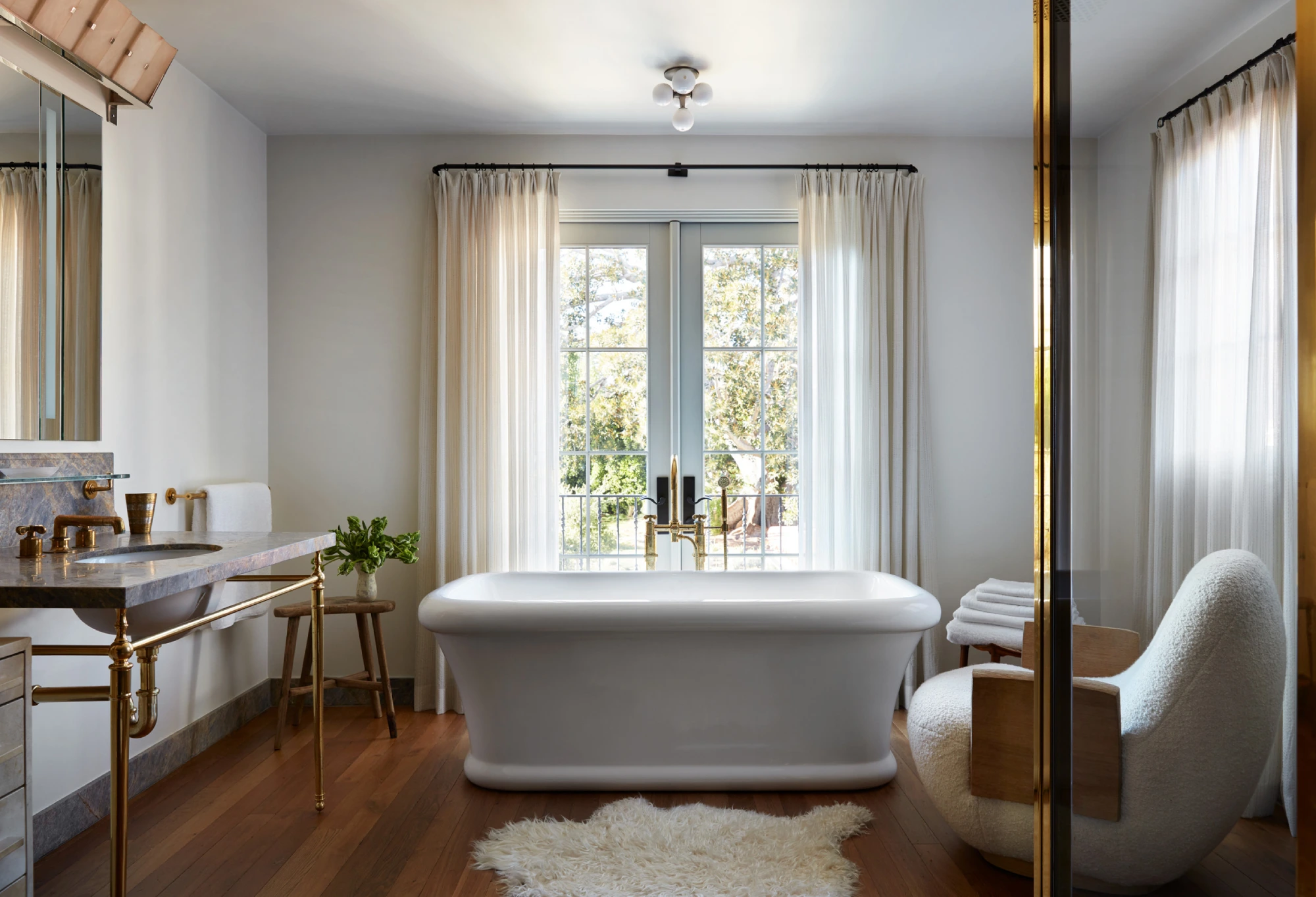 Elegant bathroom with a central white bathtub, wooden floor, and plush chair. Large window with sheer curtains lets in natural light, creating a serene ambiance.