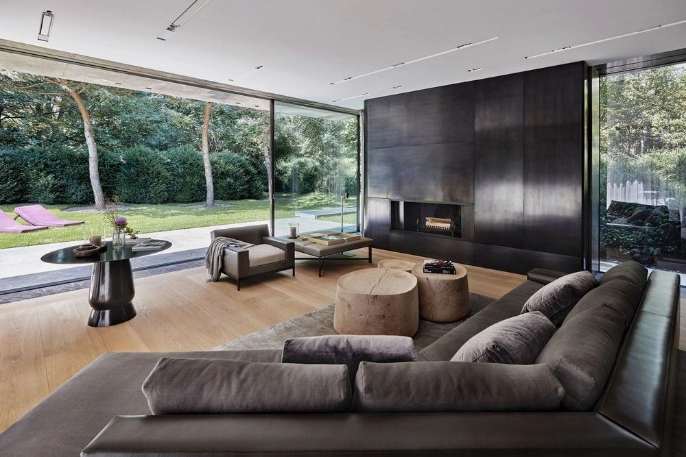 Modern living room with large gray sectional sofa facing a sleek, black fireplace. Floor-to-ceiling windows open to a lush garden, creating an airy, serene atmosphere.