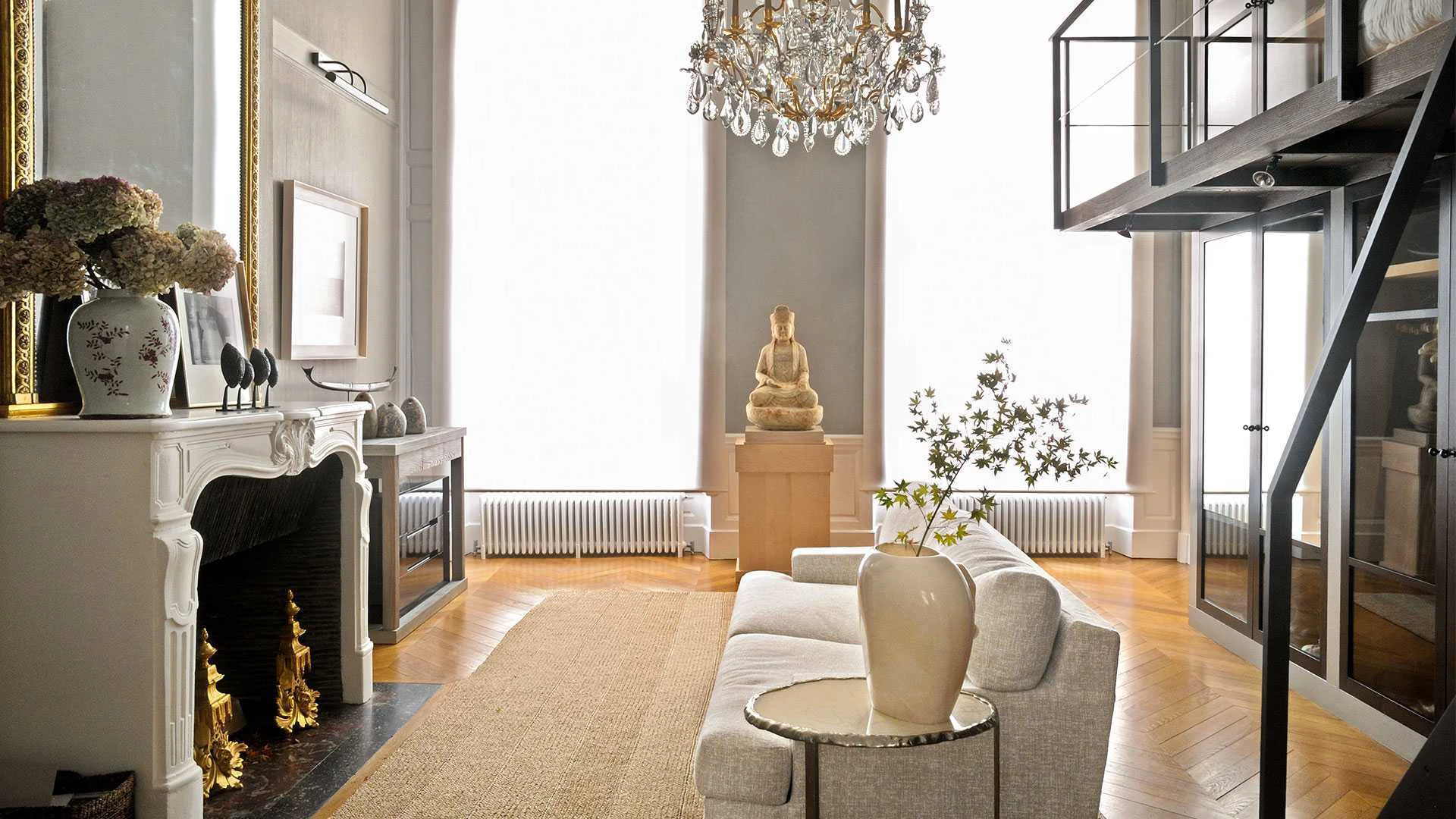 Elegant living room with chandelier, ornate fireplace, and neutral tones. A statue on a pedestal and a large vase with flowers create a serene atmosphere.