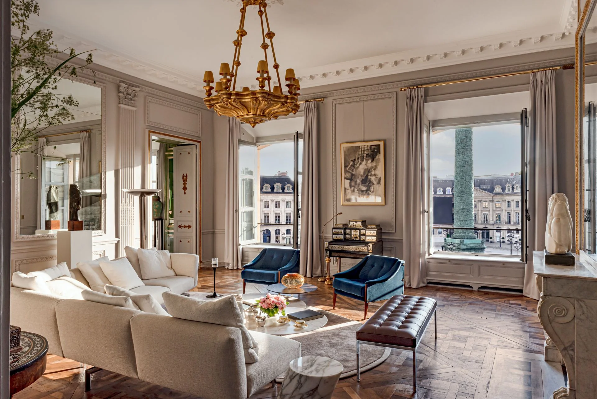 Elegant living room in Paris with ornate chandelier, beige sofas, blue chairs, and view of a historic square through tall windows. Chic, sophisticated ambiance, common of Parisian luxury apartments.