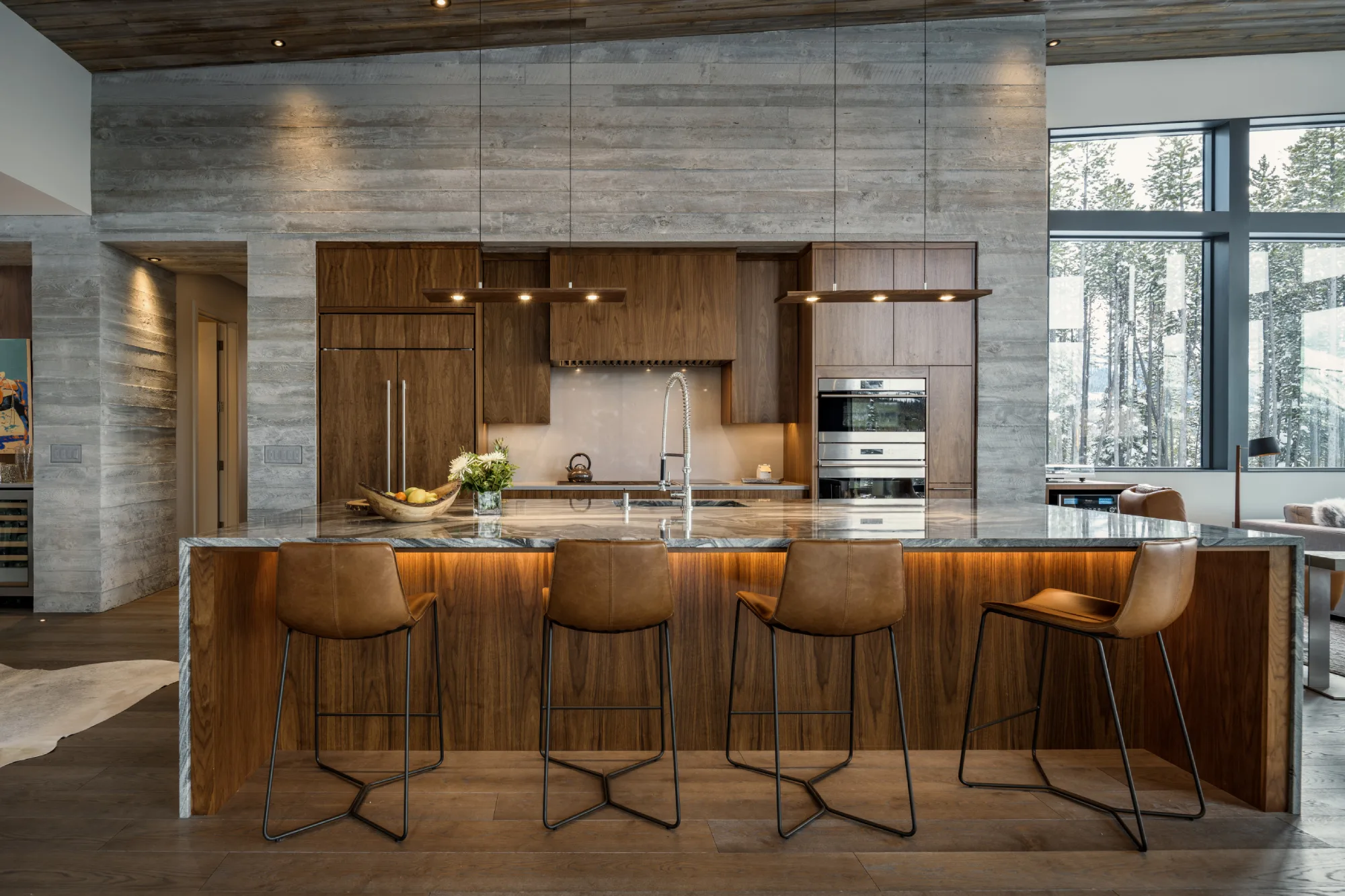 Modern kitchen with wooden cabinets and a marble countertop island, featuring four brown leather barstools. Large windows showcase a snowy forest view.