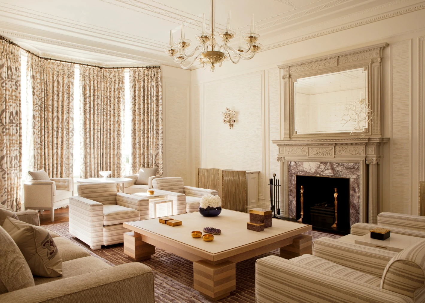 Elegant living room with cream tones, featuring a large chandelier, ornate fireplace, striped sofas, wooden coffee table, and patterned curtains. Luxurious and serene ambiance.