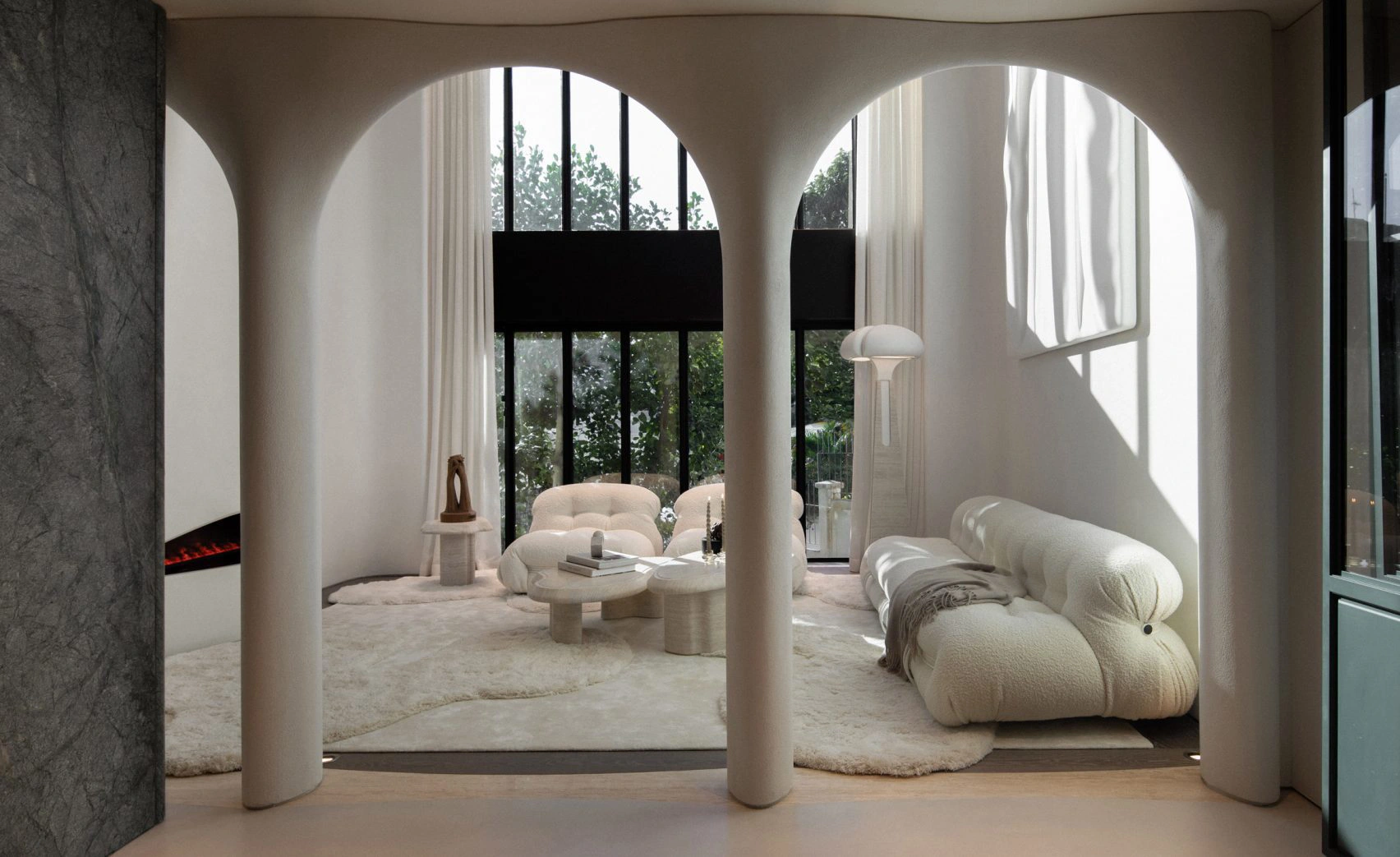 Luxurious living room with arched pillars, plush white sofas, and a cozy, modern fireplace. Soft natural light streams through large glass windows.