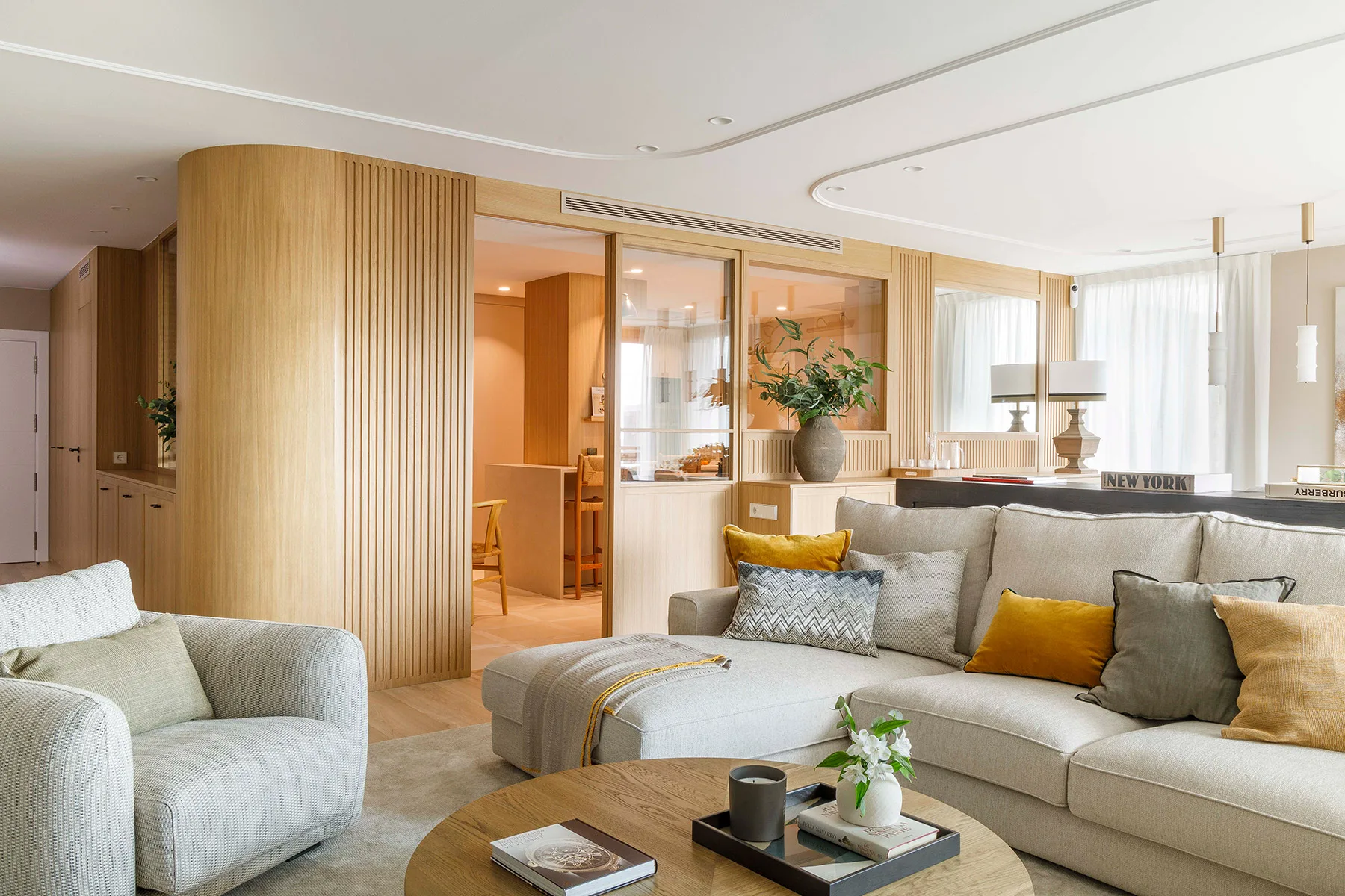 Modern living room with a cozy white sectional sofa adorned with colorful cushions, a round wooden coffee table, and soft natural light filtering through large windows. The room exudes warmth with light wood paneling and green plants, commonly found in Barcelona penthouses.