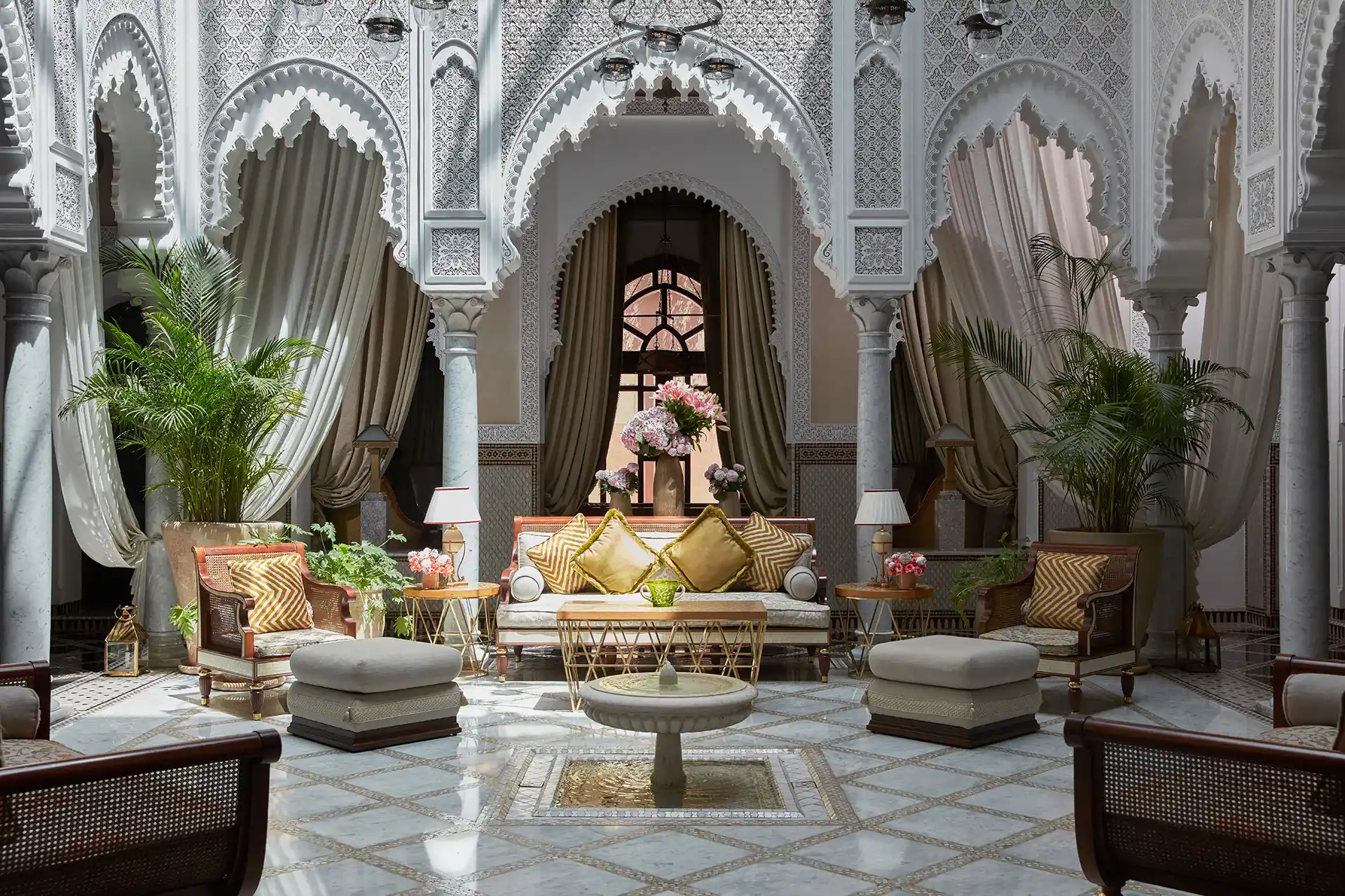 Luxurious Moroccan interior with arched doorways, intricate patterns, and plush seating. Richly decorated with plants, soft lighting, and elegant drapery, commonly found in Marrakech Riads.