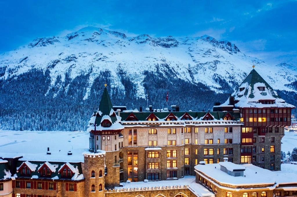 Badrutt’s Palace Hotel with illuminated windows and snow-covered turrets stands amidst a snowy landscape, framed by majestic mountains under a twilight sky.