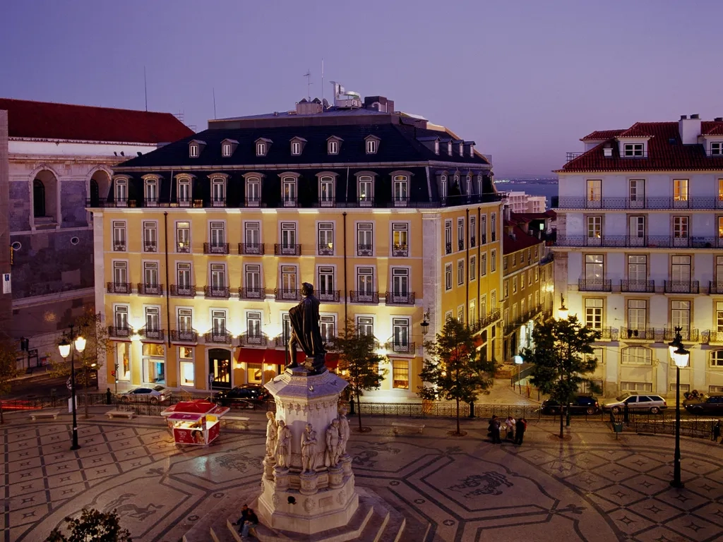 Evening view of a lit square in Lisbon with a statue centerpiece, surrounded by illuminated classic architecture, showcasing the Bairro Alto Hotel.